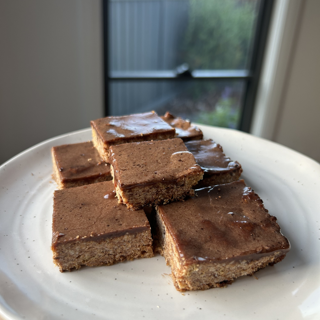 Six squares of Weet-Bix slice with a glossy chocolate topping sweetened with SaweetWay allulose, stacked on a white ceramic plate near a sunny window