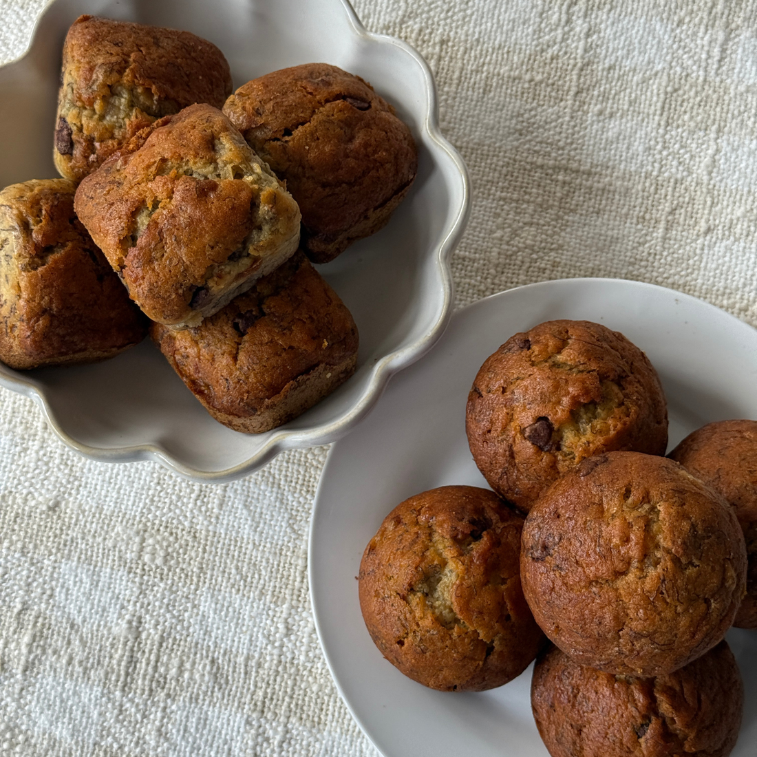 Two white ceramic dishes holding golden banana chocolate chip scones and muffins baked with SaweetWay allulose, styled on a textured linen surface