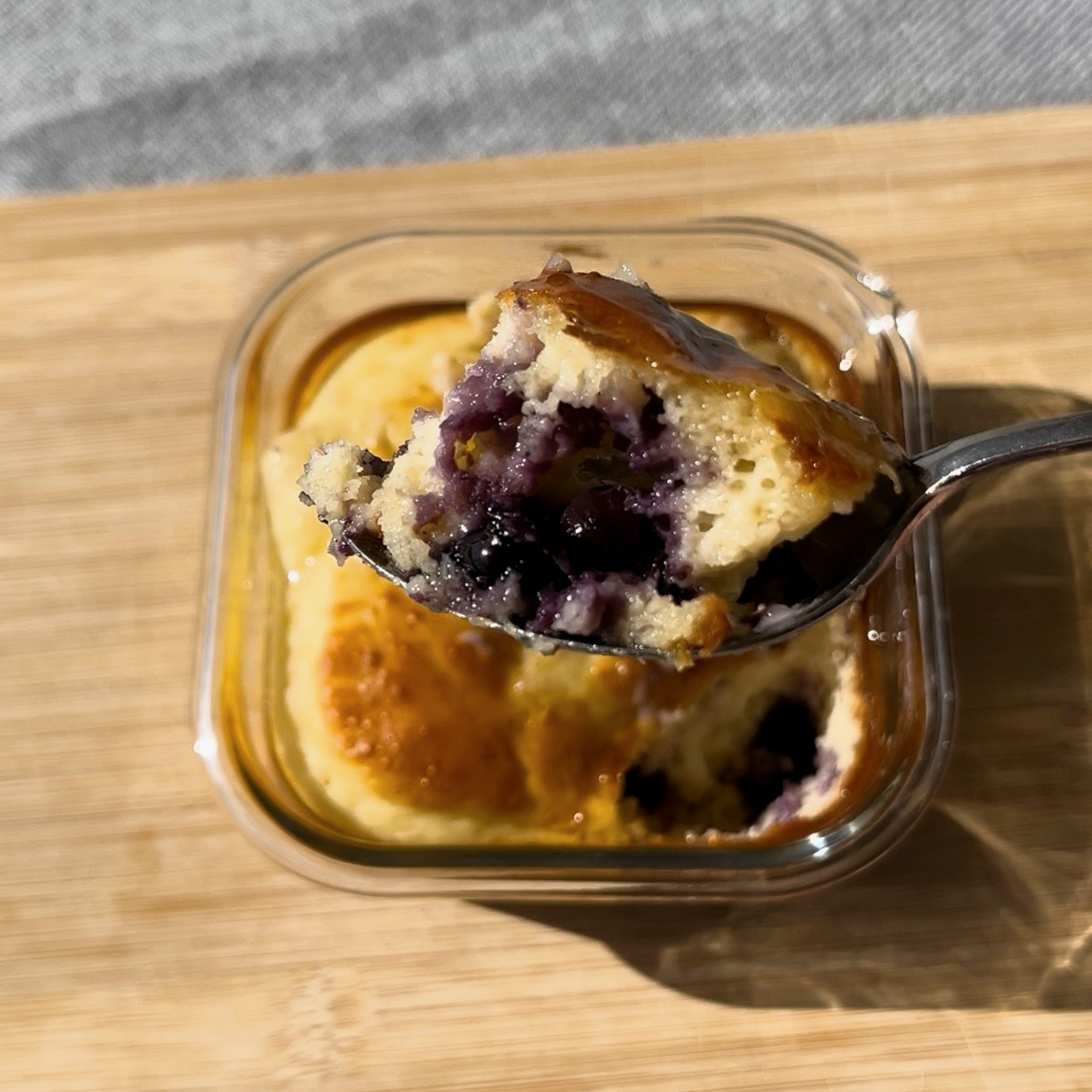 A golden blueberry mug cake baked in a square glass container on a timber board, with a spoonful lifted to reveal a fluffy crumb bursting with juicy blueberries, made with SaweetWay allulose