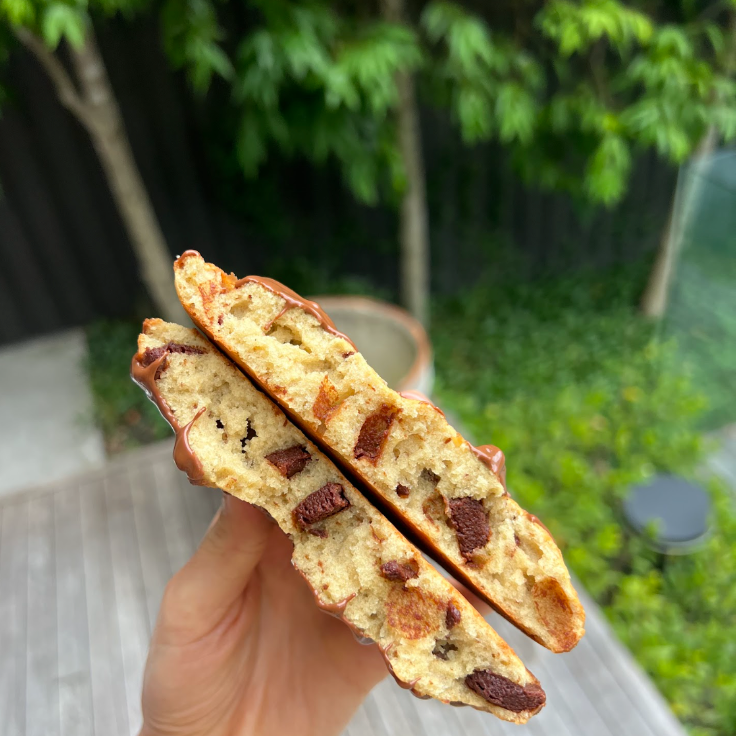 A hand holding two halves of a thick chocolate chunk cookie baked with SaweetWay allulose, showing a golden chewy crumb studded with dark chocolate pieces, with lush greenery in the background