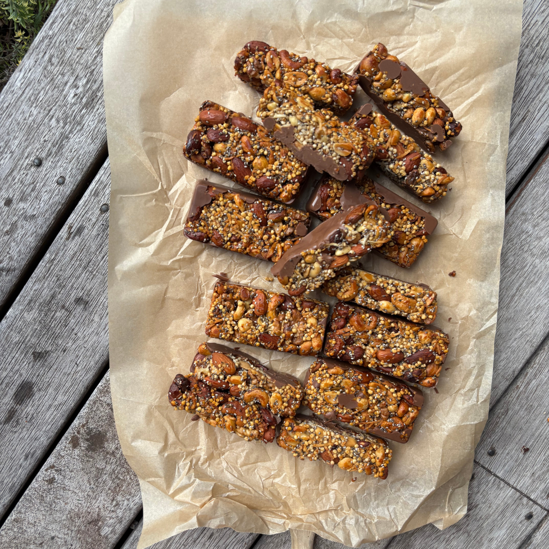 A wooden serving board lined with baking paper holding rows of chocolate-drizzled nut and seed granola bars made with SaweetWay allulose, styled on a rustic outdoor timber table