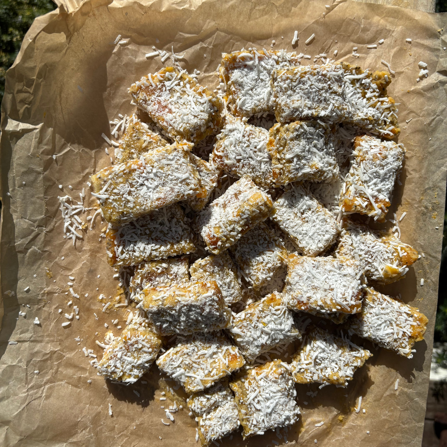 A rustic wooden board lined with baking paper holding golden lemon slice pieces generously coated in shredded coconut, made with SaweetWay allulose, styled outdoors in natural sunlight