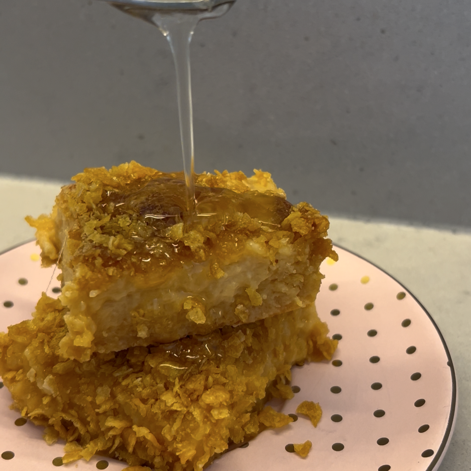 Two stacked golden cornflake slice squares on a pink polka dot plate, with a stream of SaweetWay allulose syrup being drizzled over the top