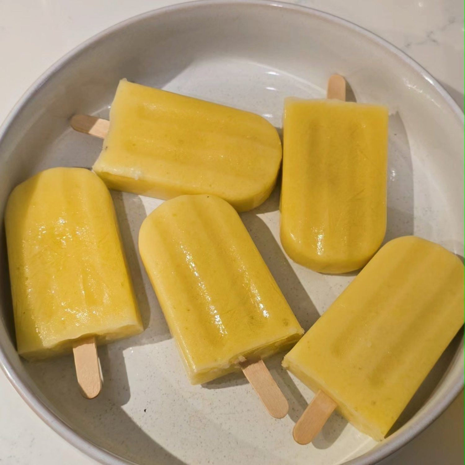 Five golden mango ice blocks made with SaweetWay allulose arranged on a round ceramic plate, smooth and glossy with wooden paddle pop sticks