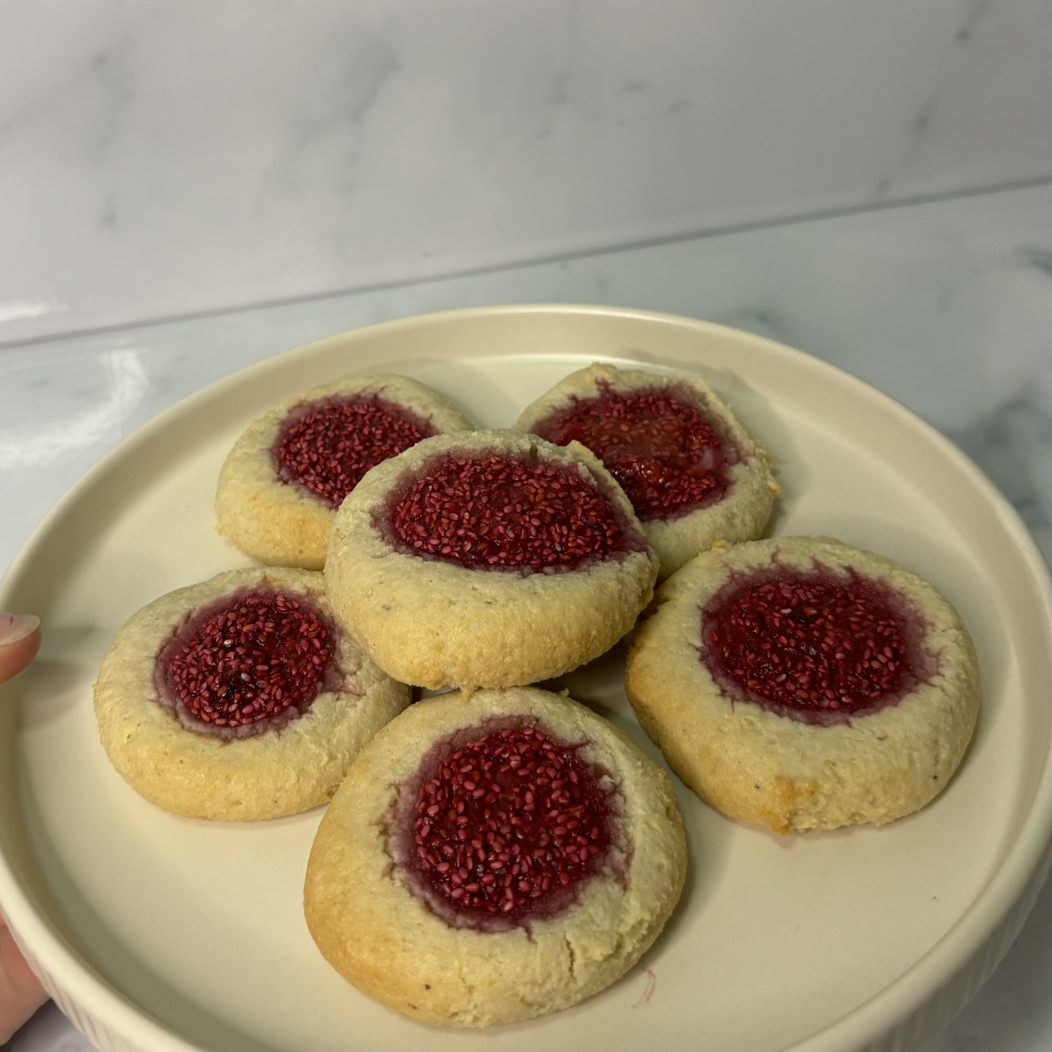 Six golden thumbprint cookies filled with raspberry chia jam made with SaweetWay allulose, arranged on a round white ceramic plate on a marble surface