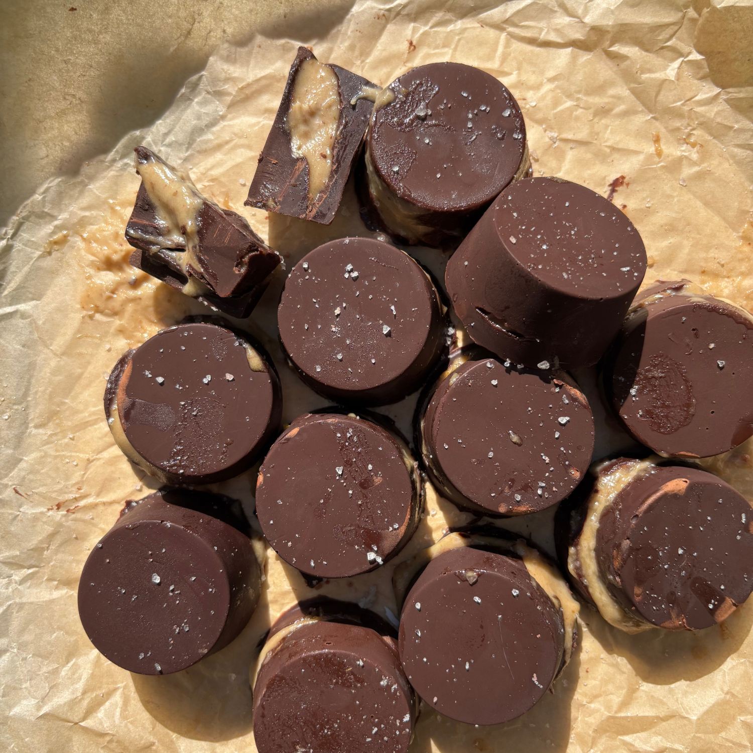 A cluster of dark chocolate salted caramel cups made with SaweetWay allulose, topped with flaked sea salt and arranged on baking paper, with one broken open to reveal the creamy caramel centre
