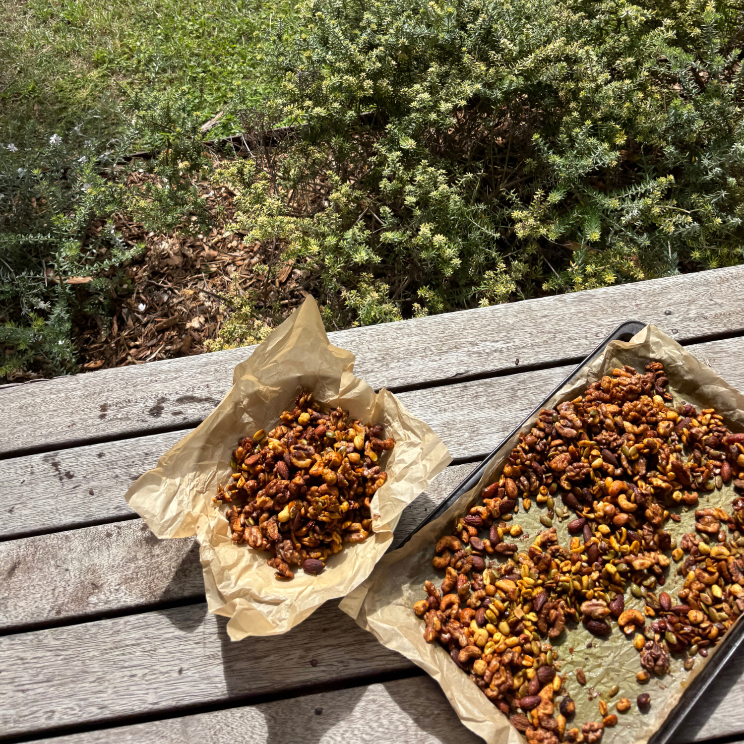 A baking tray and baking paper pile of golden spiced roasted mixed nuts made with SaweetWay allulose, cooling on a rustic outdoor timber table in the sunshine