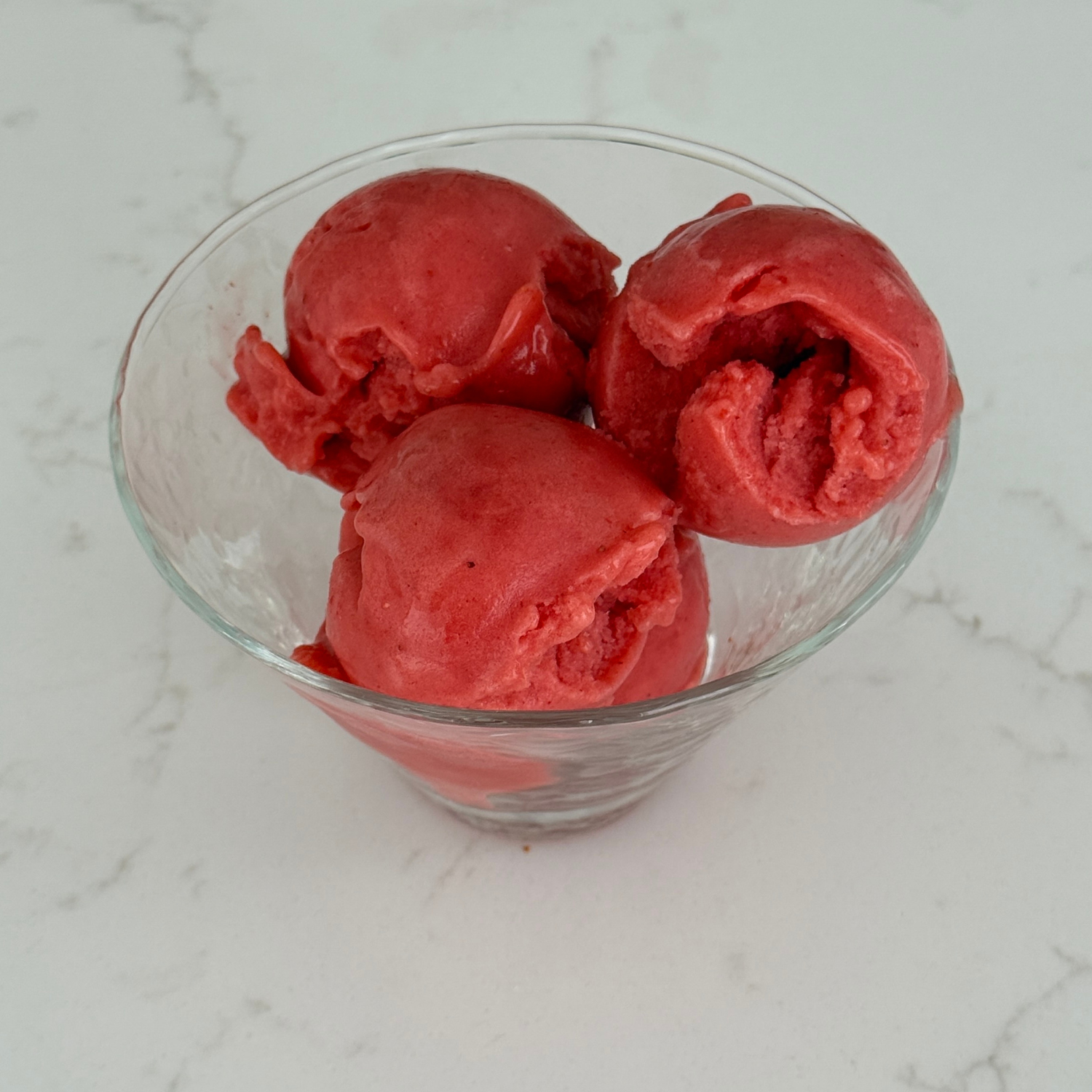 Three scoops of vibrant strawberry sorbet made with SaweetWay allulose served in a clear glass cup